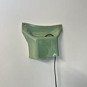May include: A wall-mounted ceramic light fixture in a light green speckled glaze. The piece features a flared, concave top section with two mounting holes and a boxy lower housing that holds a light bulb, connected by a black electrical cord.