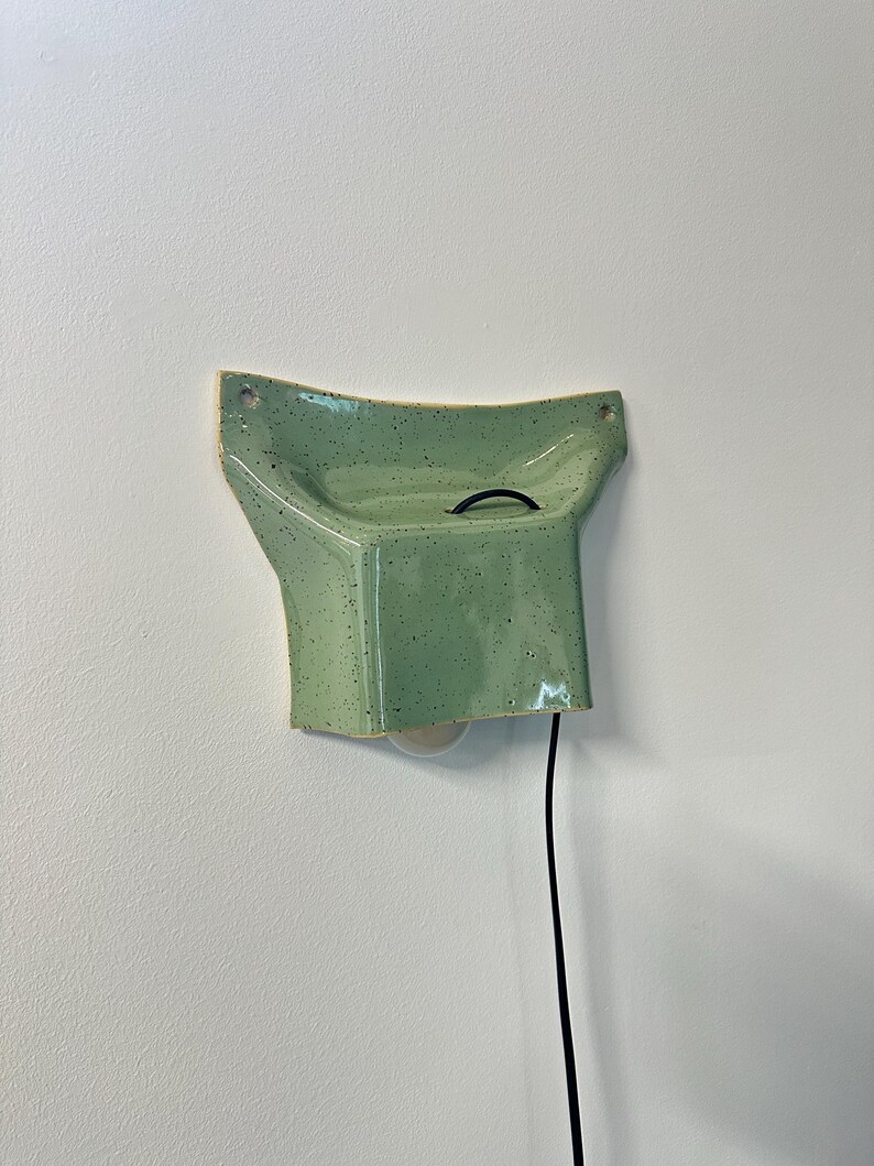 May include: A wall-mounted ceramic light fixture in a light green speckled glaze. The piece features a flared, concave top section with two mounting holes and a boxy lower housing that holds a light bulb, connected by a black electrical cord.