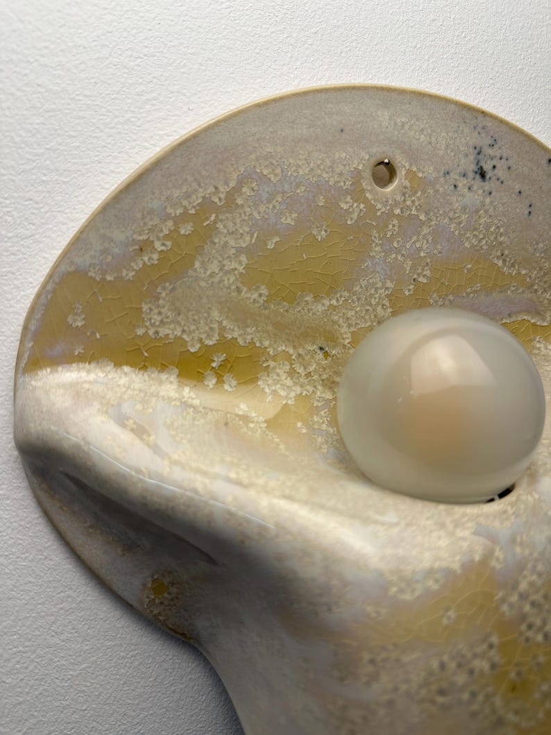May include: A wall-mounted ceramic light fixture with a semi-circular, organic shape. The surface features a crystalline glaze in shades of yellow, cream, and white with a fine crackle pattern. A single round, frosted white light bulb is attached.