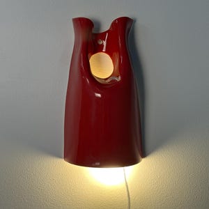 May include: A glossy red wall sconce with a smooth, curved, elongated form. The fixture features a circular cutout near the top and an open bottom that emits a warm, bright light downward, casting a glow against the wall surface behind it.