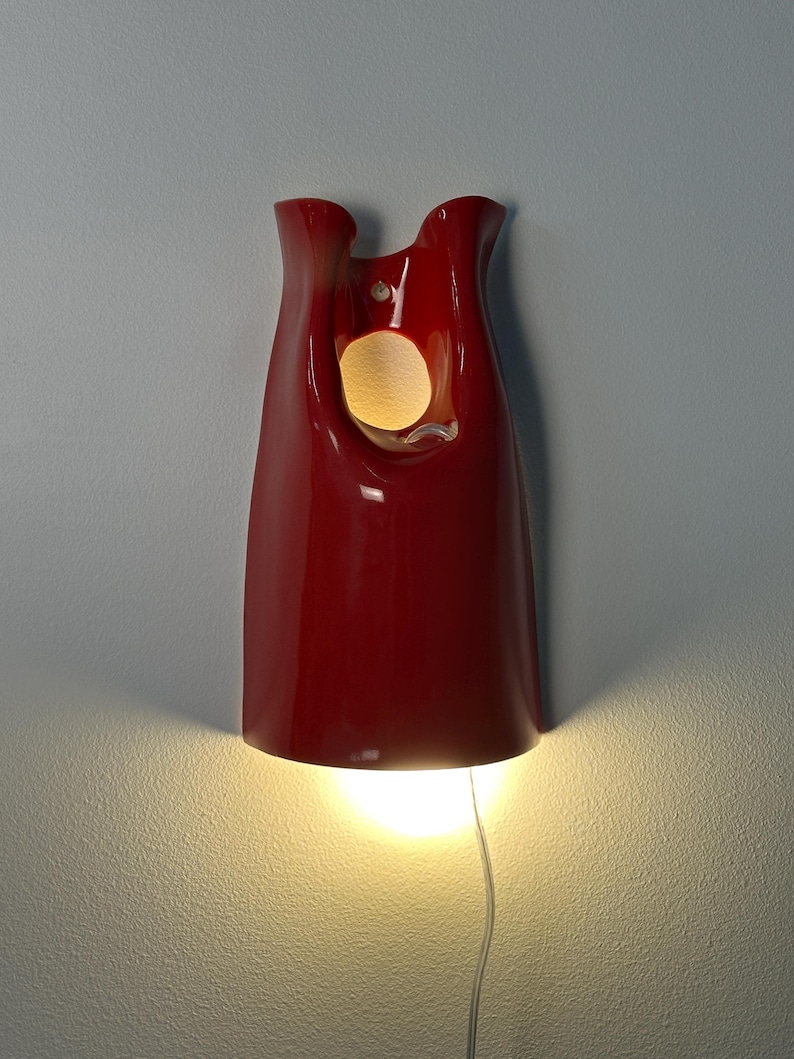May include: A glossy red wall sconce with a smooth, curved, elongated form. The fixture features a circular cutout near the top and an open bottom that emits a warm, bright light downward, casting a glow against the wall surface behind it.