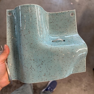 May include: A light blue ceramic wall-mounted shelf with a speckled brown pattern. The piece features a curved, wave-like design, two circular holes at the top corners for mounting, and a small metal ring centered on the flat surface area.