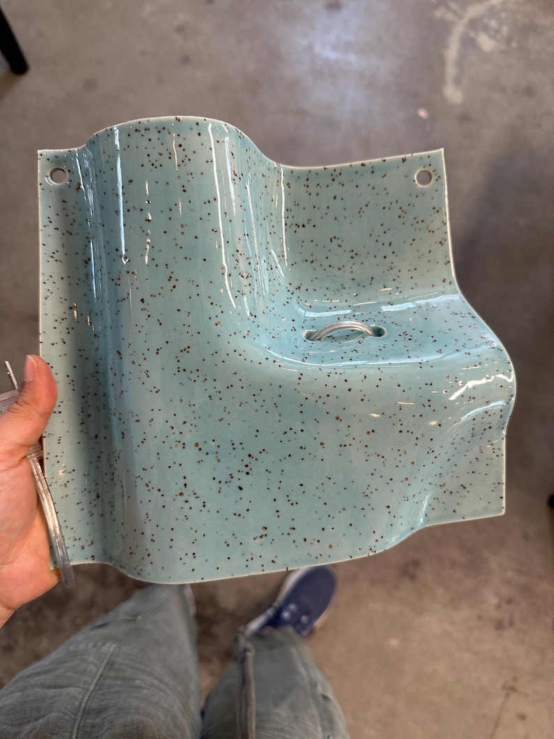 May include: A light blue ceramic wall-mounted shelf with a speckled brown pattern. The piece features a curved, wave-like design, two circular holes at the top corners for mounting, and a small metal ring centered on the flat surface area.