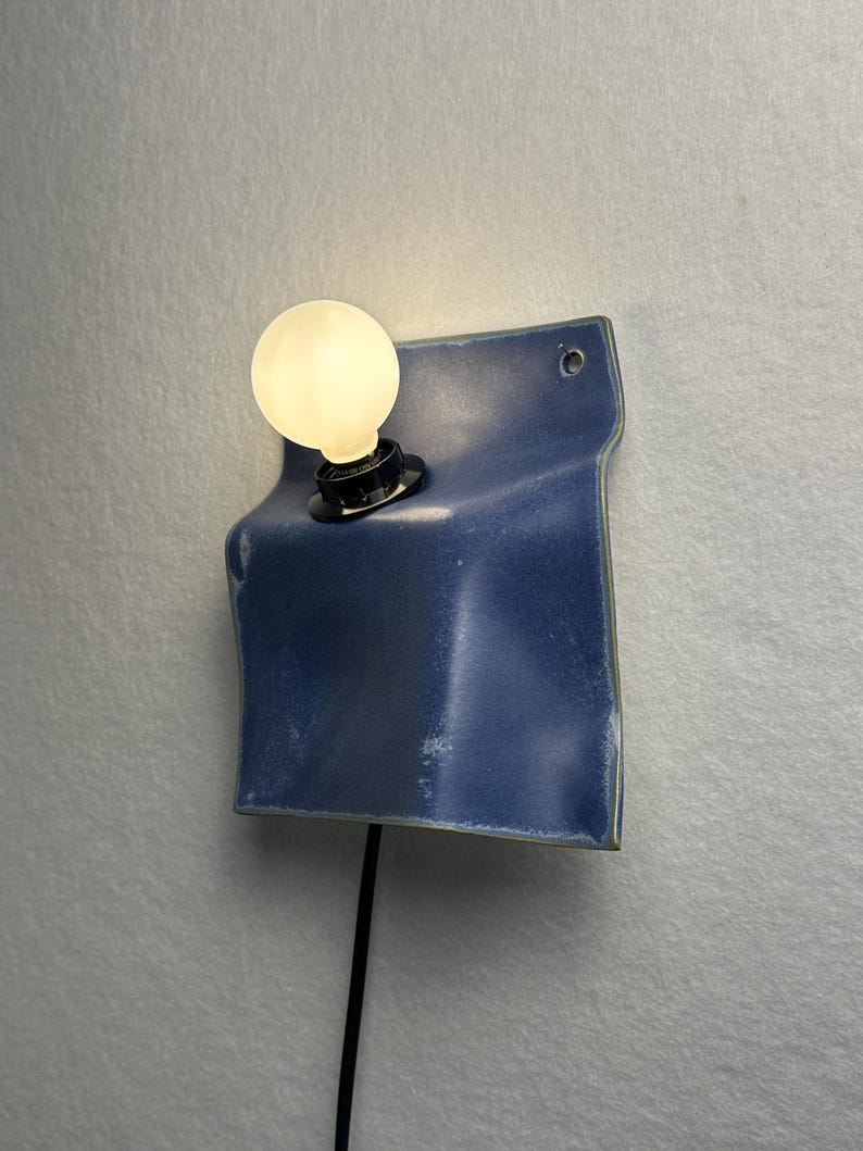 May include: A wall-mounted lamp featuring a folded, deep blue ceramic square plate with a smooth, glossy finish. A black socket holds a single, glowing round white light bulb. A thin black power cord extends downward from the base of the fixture.