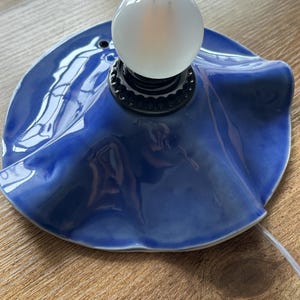 May include: A ceramic table lamp with a glossy, deep blue shade. The shade has a ruffled edge and supports a white, spherical light bulb. The lamp sits on a wooden surface, with a white cord extending from the base.