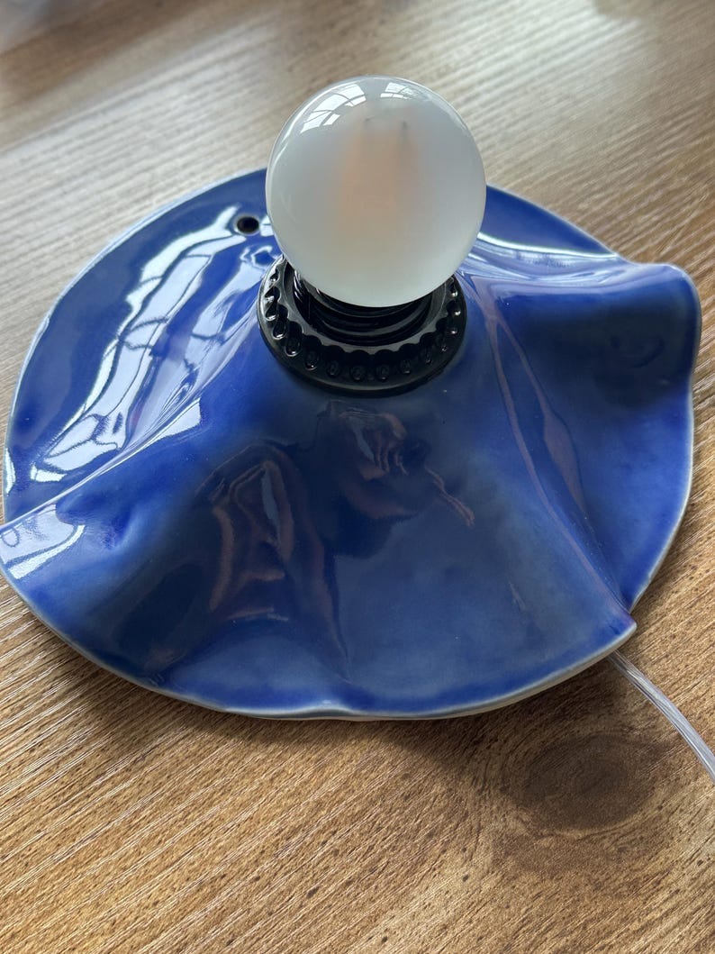 May include: A ceramic table lamp with a glossy, deep blue shade. The shade has a ruffled edge and supports a white, spherical light bulb. The lamp sits on a wooden surface, with a white cord extending from the base.