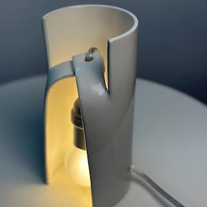 May include: A white ceramic table lamp with a cylindrical, open-sided structure. The lamp features a curved, overlapping design that creates a vertical opening, revealing a single glowing light bulb suspended from a cord inside the hollow center.