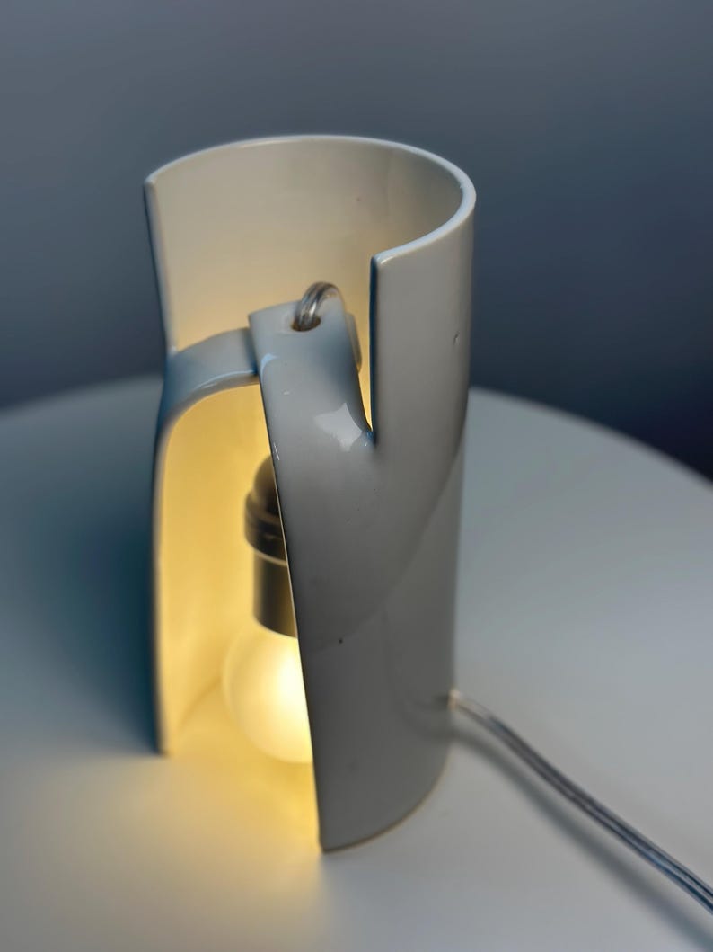May include: A white ceramic table lamp with a cylindrical, open-sided structure. The lamp features a curved, overlapping design that creates a vertical opening, revealing a single glowing light bulb suspended from a cord inside the hollow center.