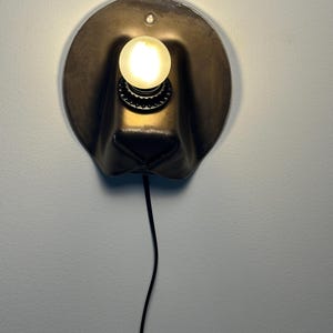 May include: A wall-mounted lamp featuring a dark, rounded, hat-shaped metal shade with a central circular opening. A single glowing, spherical light bulb is mounted in the center, and a thin black power cord extends downward from the base.