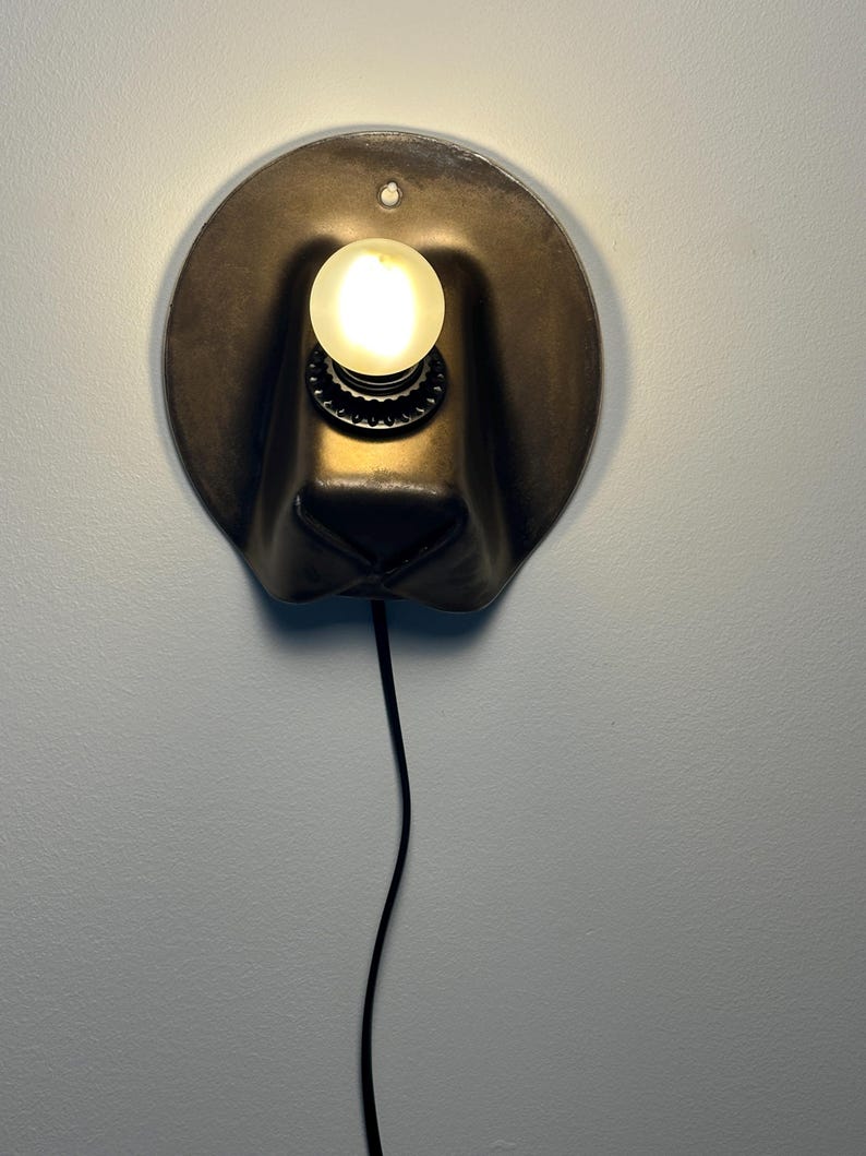 May include: A wall-mounted lamp featuring a dark, rounded, hat-shaped metal shade with a central circular opening. A single glowing, spherical light bulb is mounted in the center, and a thin black power cord extends downward from the base.