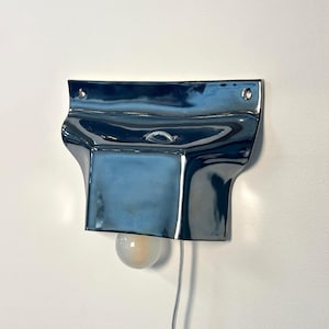 May include: A wall-mounted lamp featuring a glossy, dark blue ceramic shade with a fluid, organic shape. The fixture includes two small circular holes at the top for mounting and a single round, frosted light bulb extending from the bottom.