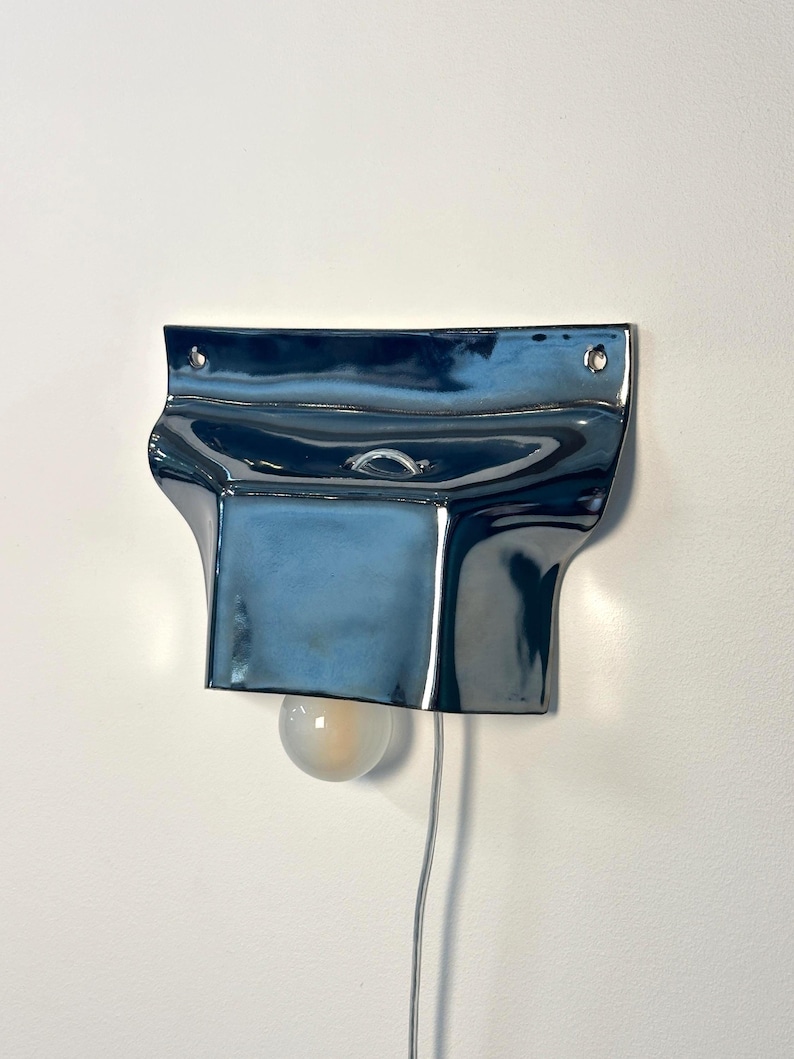 May include: A wall-mounted lamp featuring a glossy, dark blue ceramic shade with a fluid, organic shape. The fixture includes two small circular holes at the top for mounting and a single round, frosted light bulb extending from the bottom.