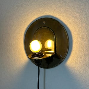 May include: A circular brass wall sconce featuring a polished reflective surface and a small, glowing spherical light bulb. The fixture includes a black power cord extending downward from the base, which is mounted flush against the wall.