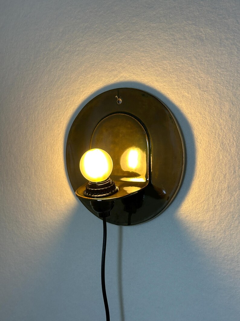 May include: A circular brass wall sconce featuring a polished reflective surface and a small, glowing spherical light bulb. The fixture includes a black power cord extending downward from the base, which is mounted flush against the wall.