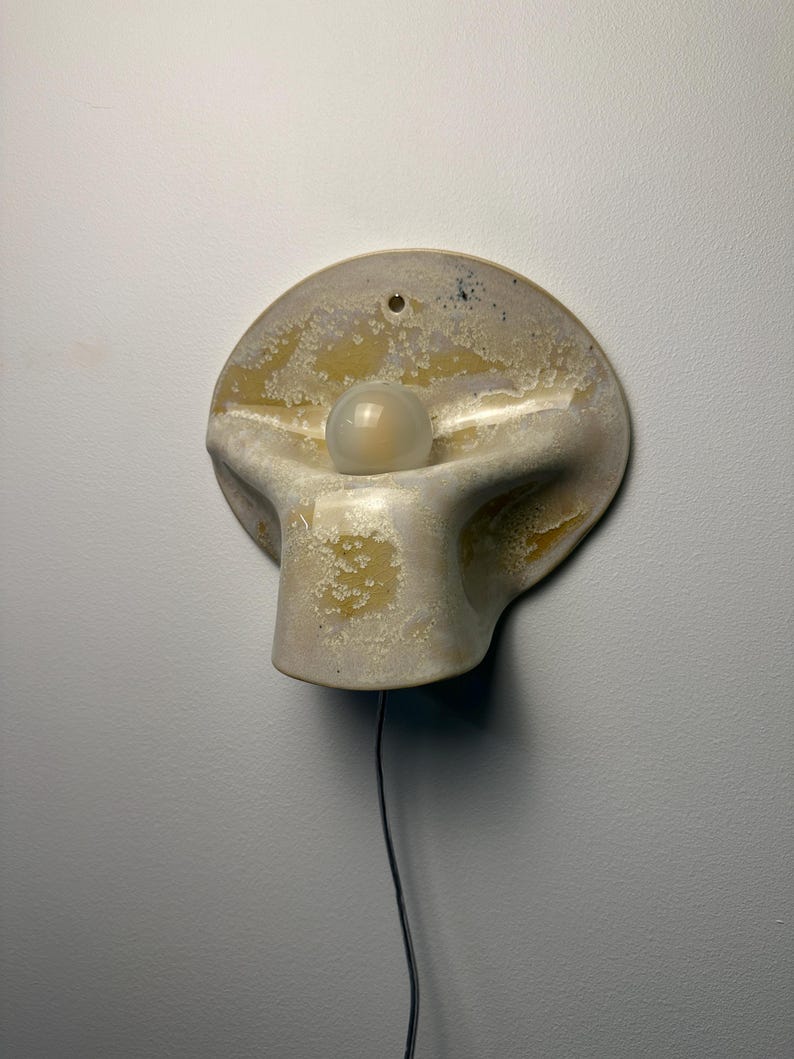 May include: A wall-mounted ceramic sconce featuring a rounded, organic shape with a mottled cream and yellow glaze. The fixture includes a central, spherical frosted glass bulb and a small hole at the top for mounting, with a black power cord extending downward.