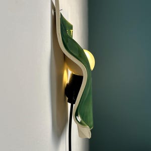 May include: A wall-mounted light fixture featuring a curved, glossy green ceramic shade with a white interior. The lamp includes a black socket and a warm, glowing light bulb, attached to a white wall with a visible nail at the top edge.