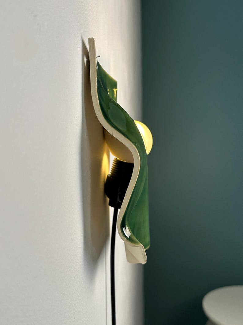 May include: A wall-mounted light fixture featuring a curved, glossy green ceramic shade with a white interior. The lamp includes a black socket and a warm, glowing light bulb, attached to a white wall with a visible nail at the top edge.