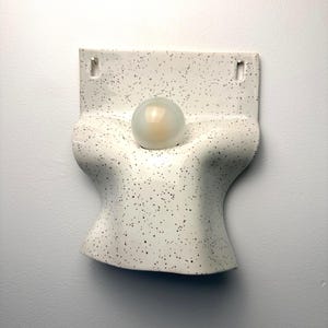 Może przedstawiać: Biały kinkiet ceramiczny w czarne kropki. Oprawa ma zakrzywiony, przypominający tuł&oacute;w kształt z centralnym okrągłym otworem, w kt&oacute;rym znajduje się pojedyncza, matowa, kulista żar&oacute;wka. U g&oacute;ry widoczne są dwa otwory montażowe do mocowania na ścianie.
