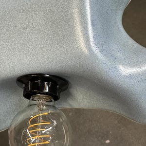 May include: A light blue ceramic lamp shade with a speckled, matte finish and an organic, wavy edge. A black socket is mounted to the shade, holding a clear glass globe light bulb with a visible, coiled yellow filament inside the bulb.