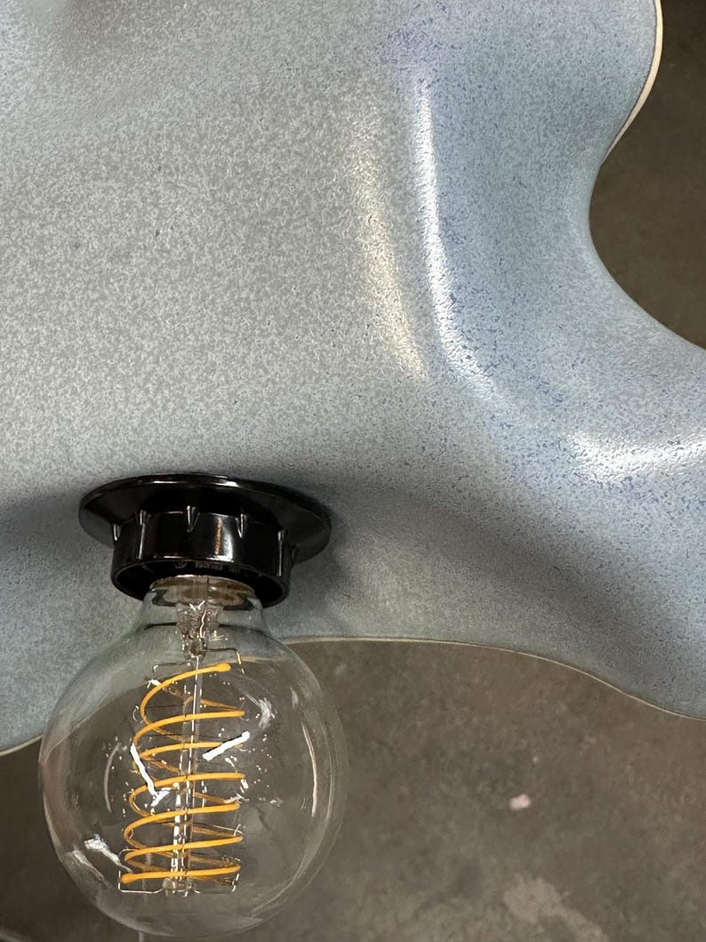 May include: A light blue ceramic lamp shade with a speckled, matte finish and an organic, wavy edge. A black socket is mounted to the shade, holding a clear glass globe light bulb with a visible, coiled yellow filament inside the bulb.