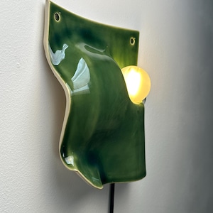 May include: A wall-mounted ceramic light fixture featuring a glossy, deep green, undulating surface with two circular mounting holes at the top. A single, warm-glowing spherical light bulb is positioned within a curved cutout on the right side.