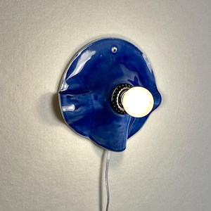 May include: A blue ceramic wall sconce with a glossy finish. The sconce has a unique, organic shape and features a light bulb in the center. A silver cord extends from the bottom. The light is on, illuminating the surrounding area.