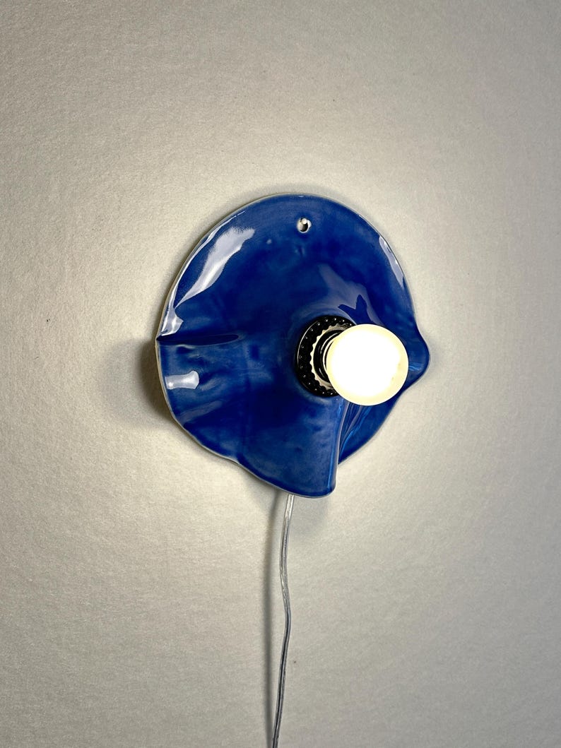 May include: A blue ceramic wall sconce with a glossy finish. The sconce has a unique, organic shape and features a light bulb in the center. A silver cord extends from the bottom. The light is on, illuminating the surrounding area.