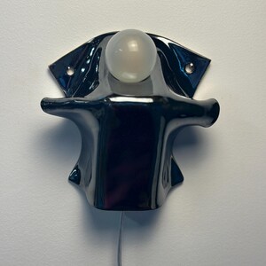 May include: A glossy black ceramic wall sconce featuring a sculptural, flared silhouette with two mounting holes on the upper wings. A single, spherical, frosted white glass light bulb is positioned at the top center, with a thin power cord extending downward.
