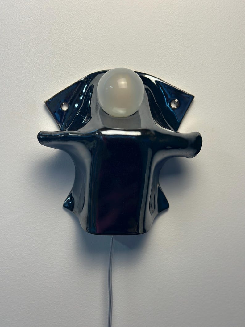 May include: A glossy black ceramic wall sconce featuring a sculptural, flared silhouette with two mounting holes on the upper wings. A single, spherical, frosted white glass light bulb is positioned at the top center, with a thin power cord extending downward.