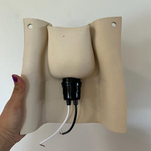 May include: A beige ceramic wall sconce featuring a curved, rectangular backplate with two mounting holes at the top. A black light socket is centred on the front, extending outward with one white wire and one black wire hanging from the base.