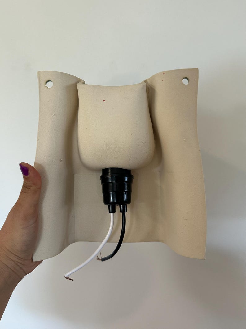 May include: A beige ceramic wall sconce featuring a curved, rectangular backplate with two mounting holes at the top. A black light socket is centred on the front, extending outward with one white wire and one black wire hanging from the base.
