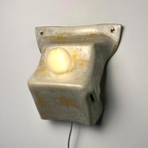 May include: A wall-mounted light fixture with a textured, off-white and yellow mottled ceramic base. The fixture features a central, glowing spherical light bulb and two small circular mounting holes at the top corners, with a thin power cord.