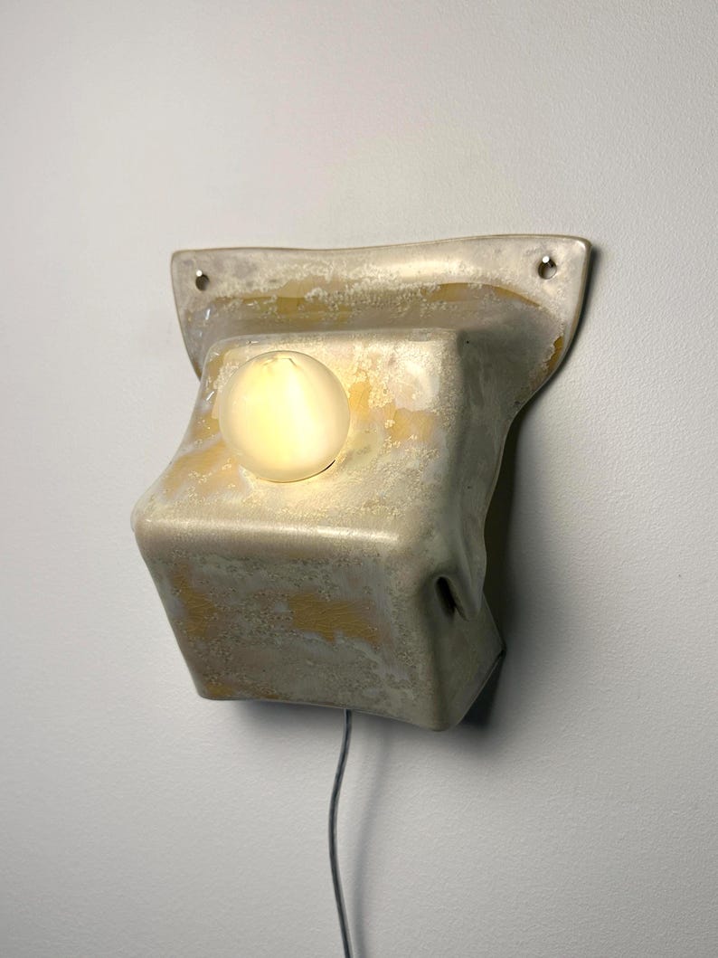May include: A wall-mounted light fixture with a textured, off-white and yellow mottled ceramic base. The fixture features a central, glowing spherical light bulb and two small circular mounting holes at the top corners, with a thin power cord.