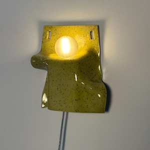 Puede incluir: Un aplique de pared de cer&aacute;mica con un esmalte brillante color chartreuse y motas negras. La pieza presenta una forma drapeada y plegada con dos orificios de montaje rectangulares en la parte superior y una bombilla blanca esf&eacute;rica encendida.