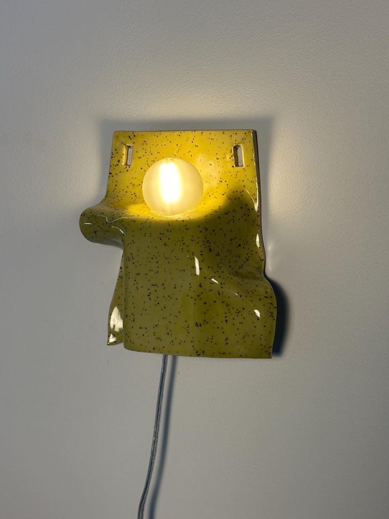 Puede incluir: Un aplique de pared de cer&aacute;mica con un esmalte brillante color chartreuse y motas negras. La pieza presenta una forma drapeada y plegada con dos orificios de montaje rectangulares en la parte superior y una bombilla blanca esf&eacute;rica encendida.