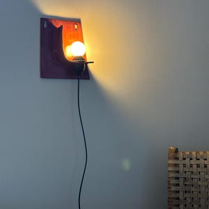 May include: A wall-mounted sconce featuring a rectangular, deep red and orange ceramic backplate with a curved shelf. A single round, glowing light bulb is seated in a black socket attached to the shelf, connected by a thin black power cord.