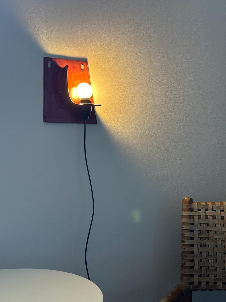 May include: A wall-mounted sconce featuring a rectangular, deep red and orange ceramic backplate with a curved shelf. A single round, glowing light bulb is seated in a black socket attached to the shelf, connected by a thin black power cord.