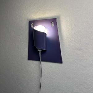 Pode incluir: Uma arandela de parede moderna com acabamento azul escuro. A arandela apresenta uma sombra curva que cobre parcialmente uma l&acirc;mpada redonda e brilhante. A lumin&aacute;ria &eacute; montada em uma placa quadrada azul escuro, com um cabo de alimenta&ccedil;&atilde;o.