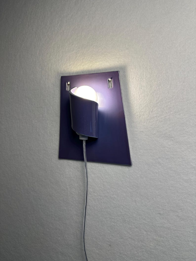 Pode incluir: Uma arandela de parede moderna com acabamento azul escuro. A arandela apresenta uma sombra curva que cobre parcialmente uma l&acirc;mpada redonda e brilhante. A lumin&aacute;ria &eacute; montada em uma placa quadrada azul escuro, com um cabo de alimenta&ccedil;&atilde;o.