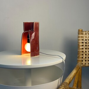 May include: A table lamp with a glossy, deep red ceramic shade featuring a curved, open-front architectural design. The lamp houses a single glowing light bulb in a black socket and includes a clear electrical cord extending from the base.