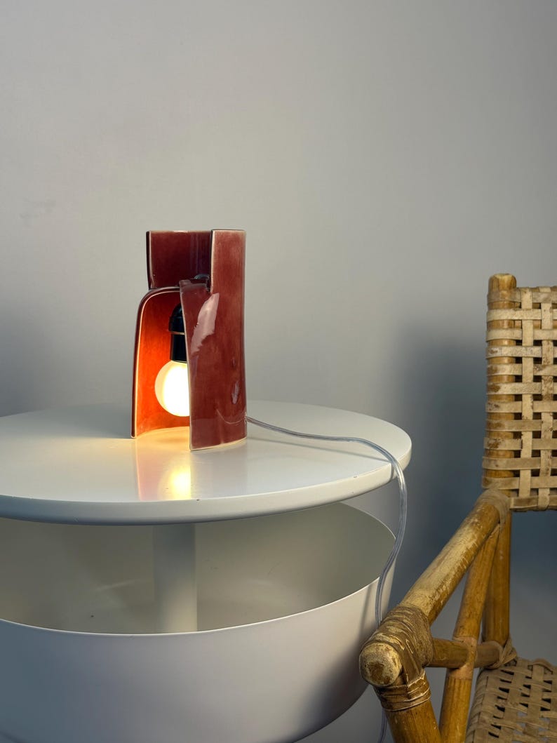 May include: A table lamp with a glossy, deep red ceramic shade featuring a curved, open-front architectural design. The lamp houses a single glowing light bulb in a black socket and includes a clear electrical cord extending from the base.