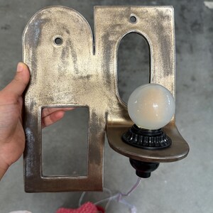 K&ouml;nnte beinhalten: Eine Wandleuchte mit metallischem, antik-bronzenem Finish, die einen bogenf&ouml;rmigen und einen rechteckigen Ausschnitt aufweist. Eine kleine, runde, mattwei&szlig;e Gl&uuml;hbirne sitzt in einer schwarzen Fassung auf einer geschwungenen Ablage.