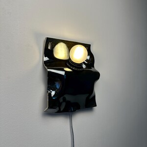 May include: A wall-mounted lamp featuring a glossy black, irregularly shaped, undulating ceramic or metal base. A single, round, frosted white glass light bulb is mounted on the upper right side, emitting a warm glow against the dark surface.