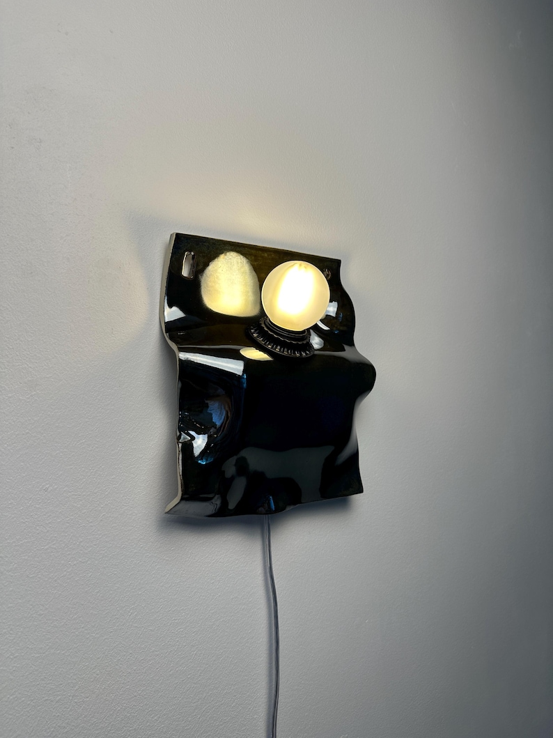 May include: A wall-mounted lamp featuring a glossy black, irregularly shaped, undulating ceramic or metal base. A single, round, frosted white glass light bulb is mounted on the upper right side, emitting a warm glow against the dark surface.