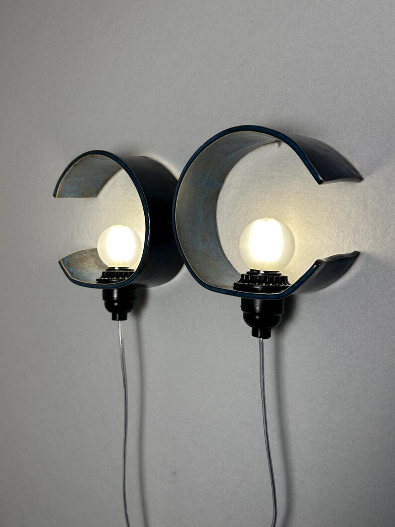 May include: Two wall-mounted lamps featuring curved, dark blue ceramic shades with a matte finish. Each lamp includes a black socket holding a round, glowing white light bulb. A thin, clear electrical cord extends downward from each lamp base.