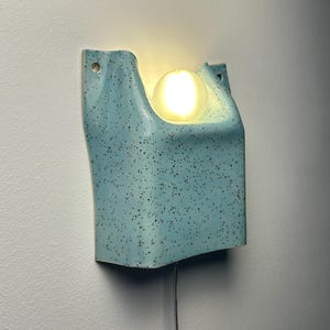 May include: A wall-mounted light fixture made of light blue ceramic with a dark speckled pattern. The fixture has a U-shaped top section that holds a single glowing, spherical white light bulb. A thin power cord extends from the bottom center.