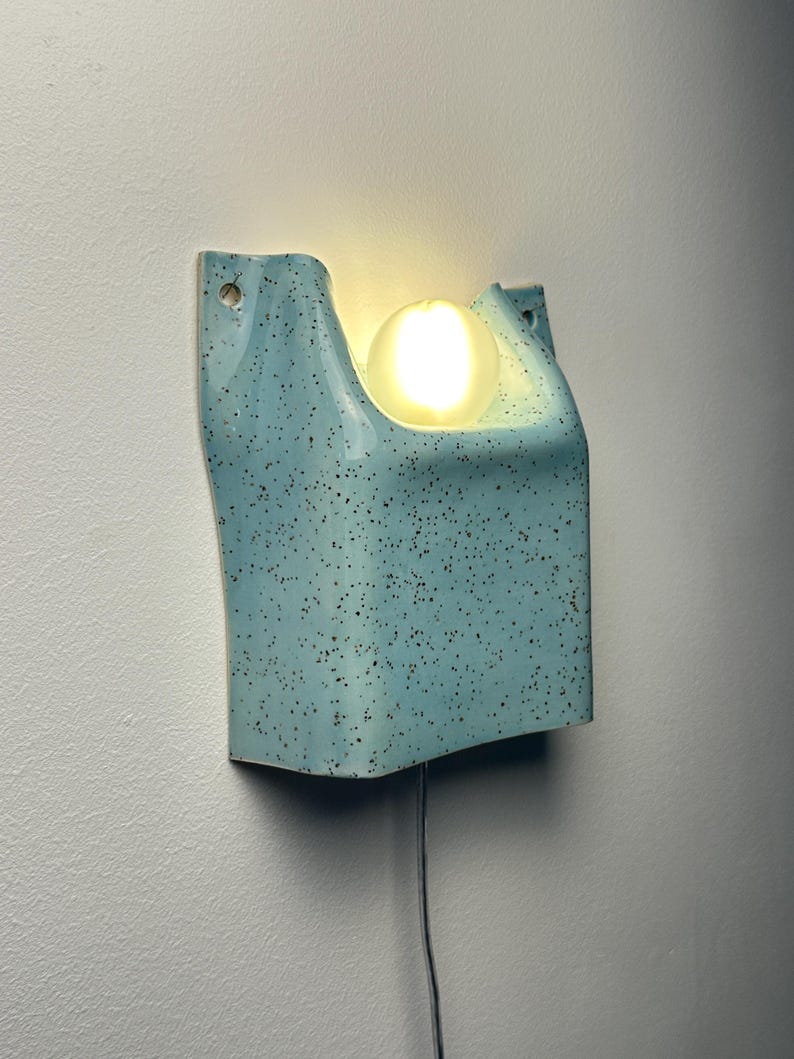 May include: A wall-mounted light fixture made of light blue ceramic with a dark speckled pattern. The fixture has a U-shaped top section that holds a single glowing, spherical white light bulb. A thin power cord extends from the bottom center.