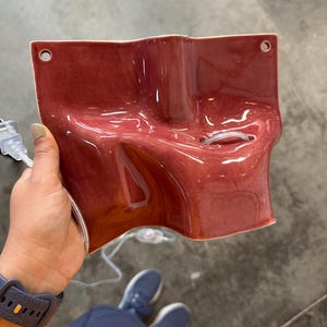 May include: A glossy, dark red ceramic wall sconce with an irregular, undulating surface. The fixture features two circular mounting holes at the top corners and a central opening for a light bulb, accompanied by a clear electrical cord and plug.