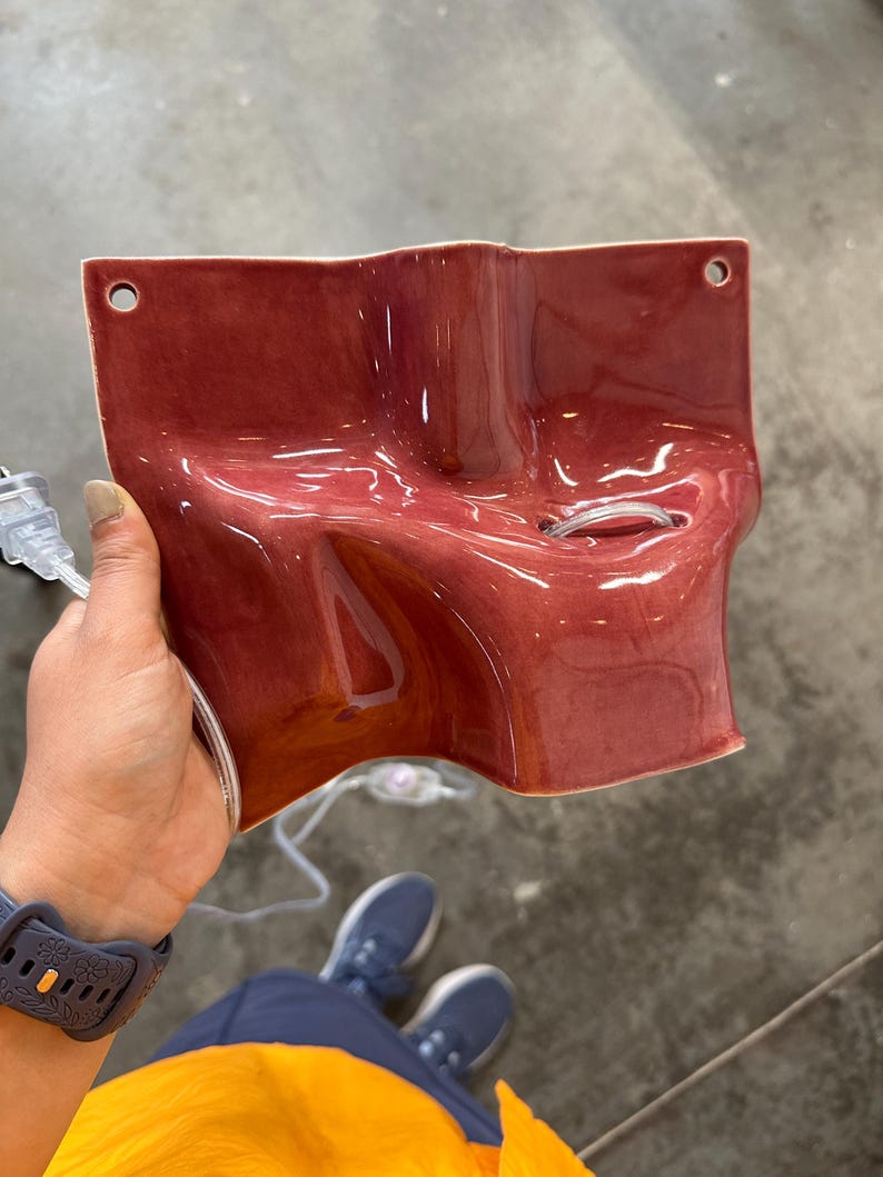 May include: A glossy, dark red ceramic wall sconce with an irregular, undulating surface. The fixture features two circular mounting holes at the top corners and a central opening for a light bulb, accompanied by a clear electrical cord and plug.