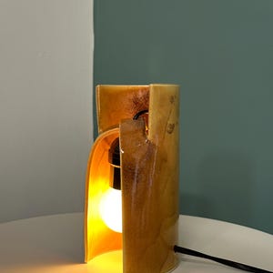 Puede incluir: Una l&aacute;mpara de mesa cil&iacute;ndrica de cer&aacute;mica con un esmalte brillante de tonos &aacute;mbar y marr&oacute;n dorado. La l&aacute;mpara presenta una abertura vertical que revela una bombilla encendida en su interior. Un cable de alimentaci&oacute;n negro sale del lateral.